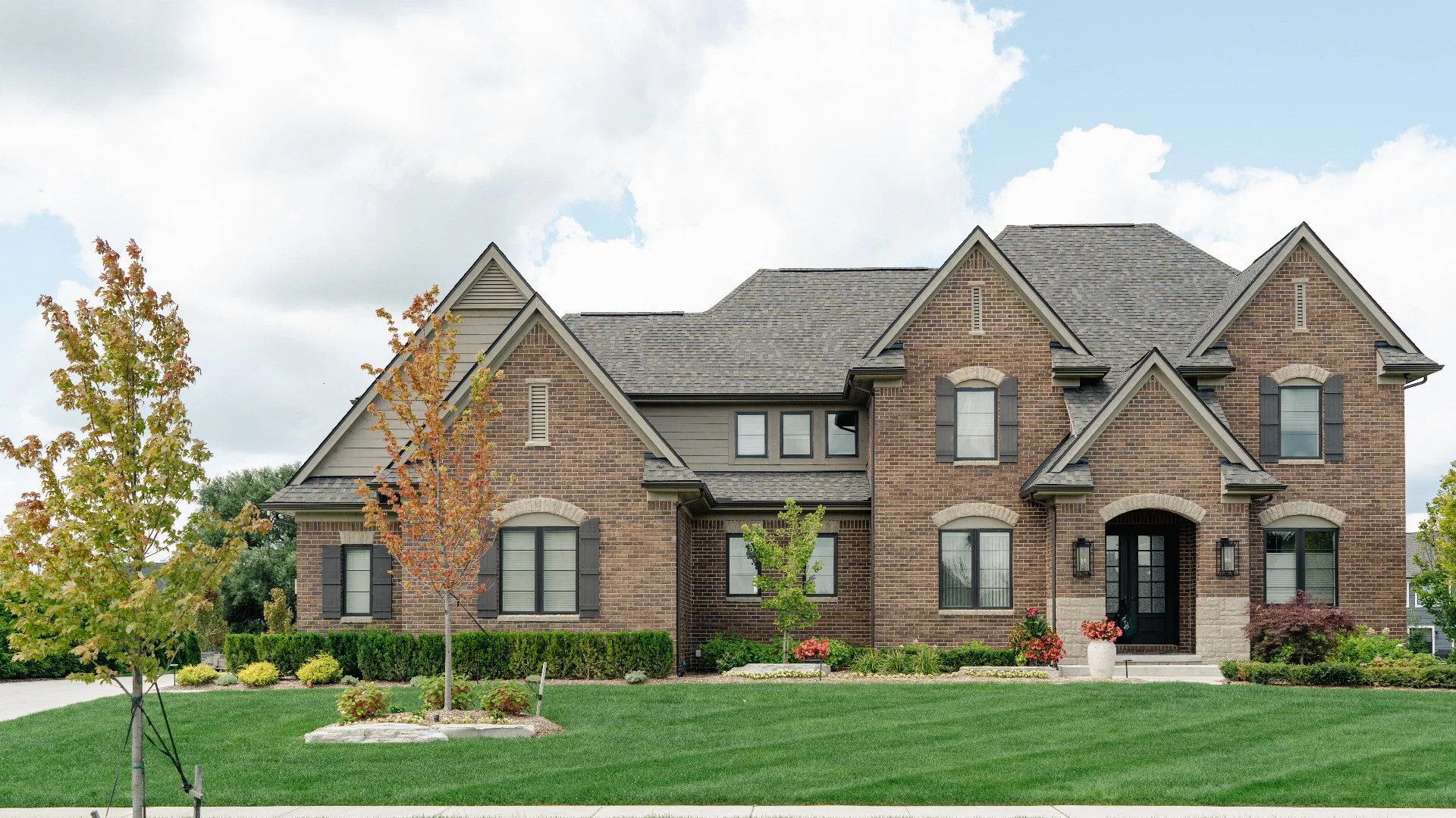 A modern elegant brick house with a well-maintained landscaped garden featuring trees and a lawn under a partly cloudy sky.