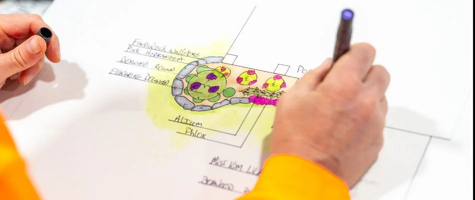 A close-up of a person's hand drawing a colorful landscape design on paper, featuring plant placements and a stone pathway, with labeled elements for planting areas.