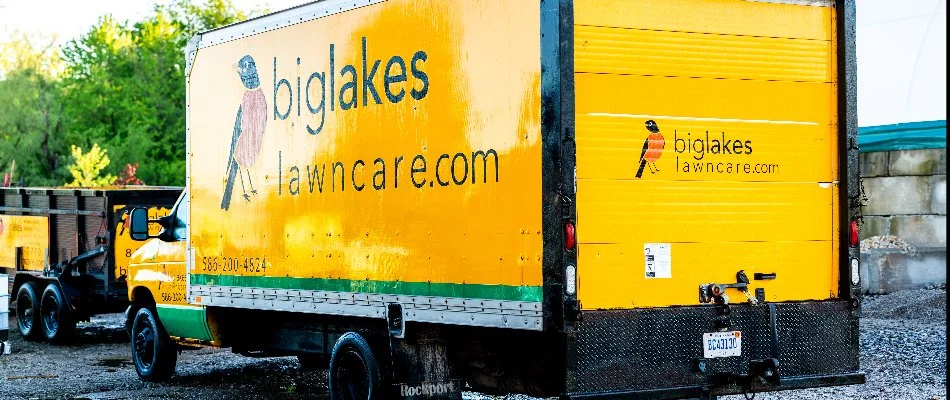 Bright yellow delivery truck with a logo for Big Lakes Lawn Care featuring contact information and a bird graphic, parked in a yard