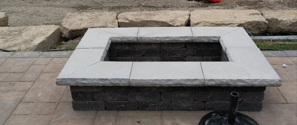 A partially constructed stone fire pit featuring a concrete top on a block base, with construction materials and safety fencing visible in the background.